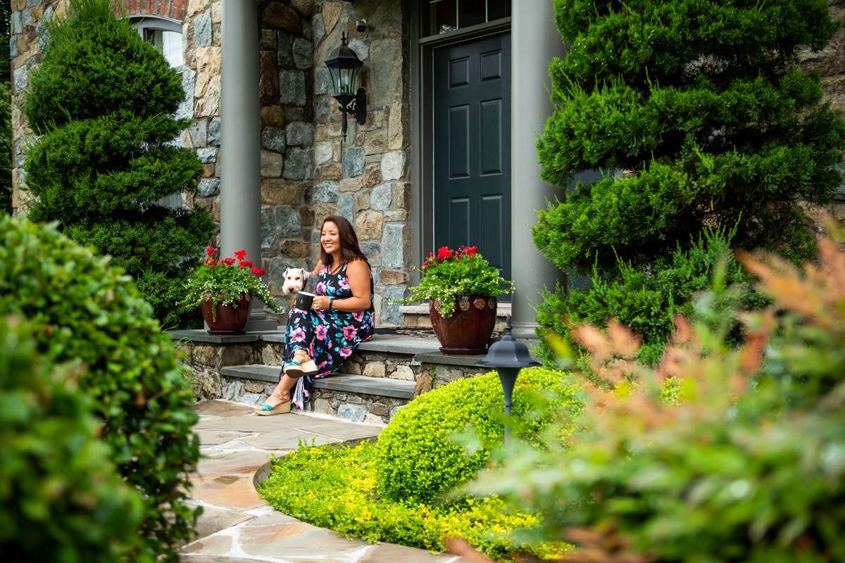 woman sits on front porch with dog with large trees and stone walkway